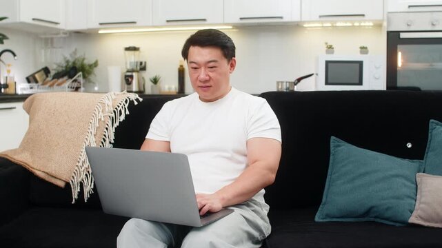 Asian man at home launches laptop boots system and begins work typing with calm smile. Chinese guy on sofa opens app checks inbox sets plan for day and starts first task then sets timer for focus