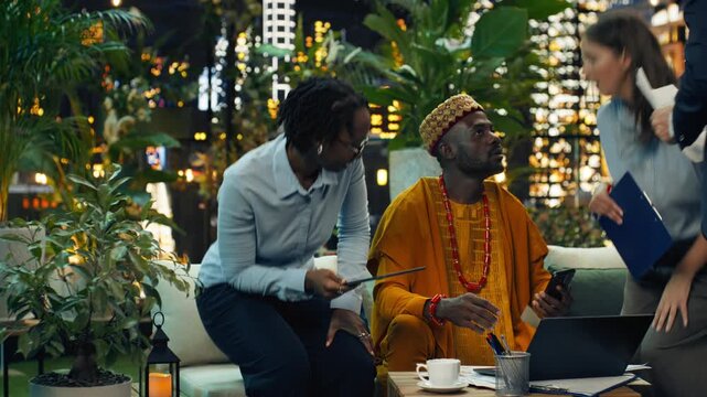 The manager in traditional attire takes a work phone call with coworkers at a rooftop workspace, where a laptop and documents support team coordination, task management and project updates.