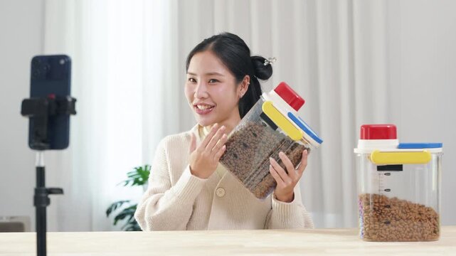 Asian woman pet food reviewer presenting airtight dog food storage container to smartphone camera at home