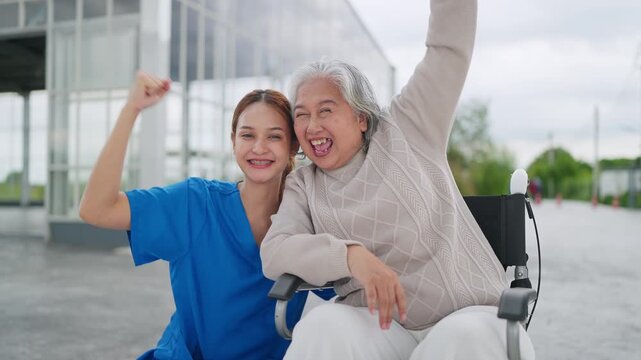 Bright smiles shine on both caregivers. Woman in blue sits beside wheelchair-bound senior. They gesture joyfully, arms raised high
