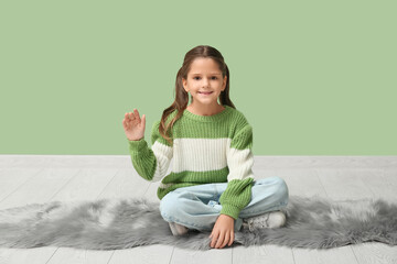 Cute little happy girl waving hand while sitting on carpet near green wall © Pixel-Shot