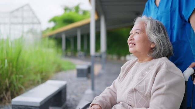 Senior woman smiles, enjoying outdoor wheelchair ride. Caregiver in blue supports her gently. Natural garden path with greenery and bench