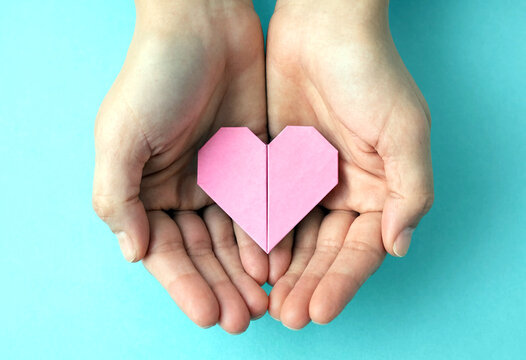 Two hands gently holding a pink paper heart symbolizing love and care against light blue background
