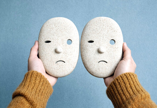 Two hands holding sad expression white ceramic masks against light blue background