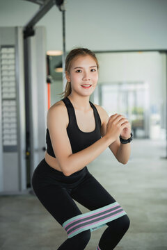 Young woman in black workout outfit exercising with resistance band around legs while squatting indoors in gym