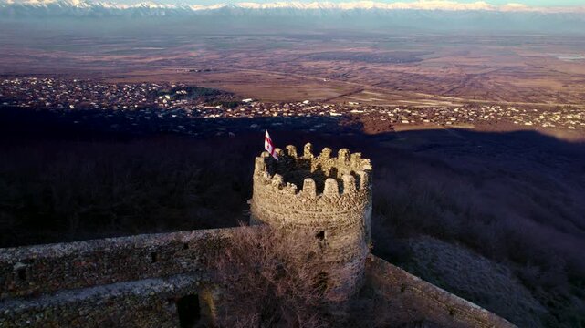Georgian flag flies from a crenellated round tower at the end of a stone wall while a town and broad farm plain spread below the wooded ridge and snowy mountains line the distant horizon