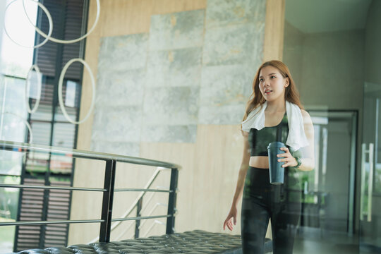 Young woman in athletic wear holding sports bottle walking indoors after workout with towel around neck in modern interior