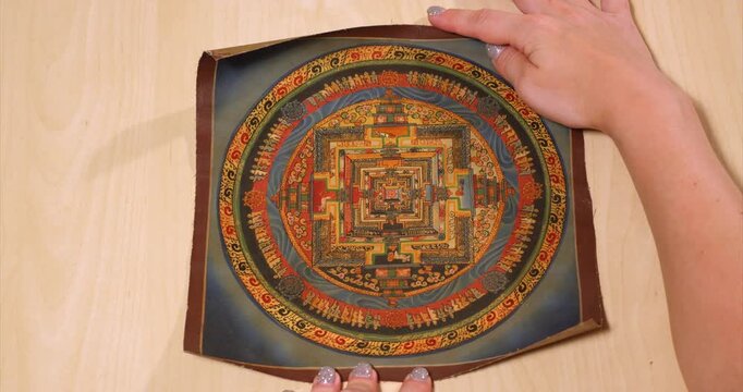 Traditional Nepalese mandala opens from a brown scroll on a wooden table revealing orange squares inside blue bands of Buddhist symbols before a hand rolls the painting closed in an overhead view