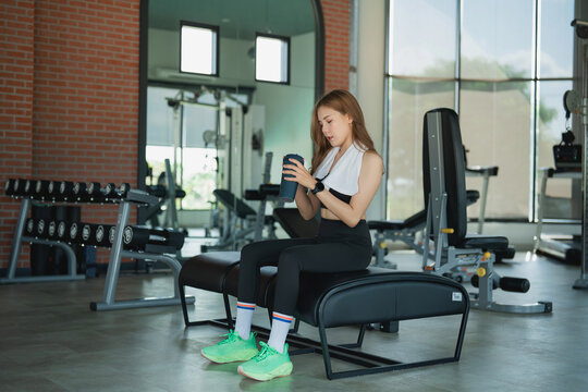 Young woman sitting on gym bench using smartphone with towel around neck in modern fitness center interior