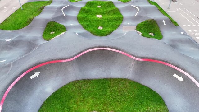 Oblique drone view across an empty asphalt pump track with rolling humps, banked curves, white direction arrows and bright grass islands forming a rhythmic geometric course.