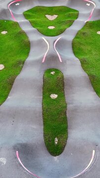 Oblique aerial view of a modern pump track with split asphalt lanes, a banked U-turn, grassy median and direction markings designed for cycling skills practice.