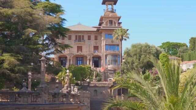 Ornate villa with a roof tower behind landscaped terraces, garden balustrades and palm fronds