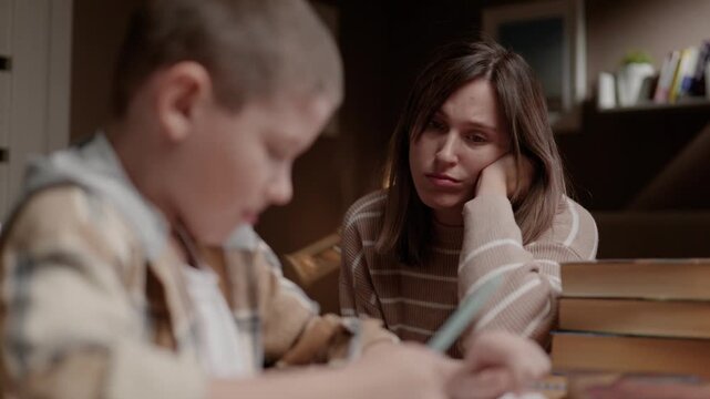 Young schoolboy doing homework at home while mother sits beside him and supervises his work. Parenting education supervision family concept