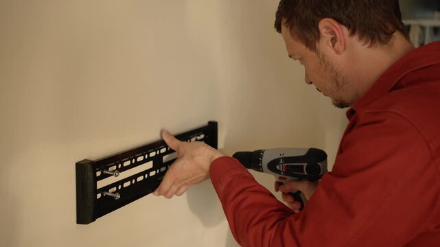 Diligent man in a red work shirt using a power drill to install a black metal bracket for a television, concentrating on the home renovation and repair task in a living room