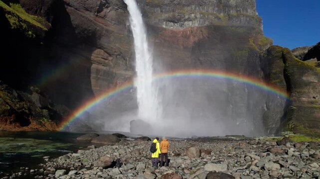Drone flies forward past two tourists in bright jackets toward Haifoss waterfall, Iceland, revealing vivid rainbow through mist above rocky riverbank, rushing river and towering volcanic canyon cliffs