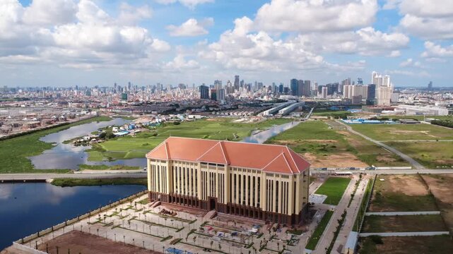 Description: Aerial view south of Phnom Penh, Cambodia, showing a large civic building beside wetlands and new roads, with rapid development and the city skyline beyond.