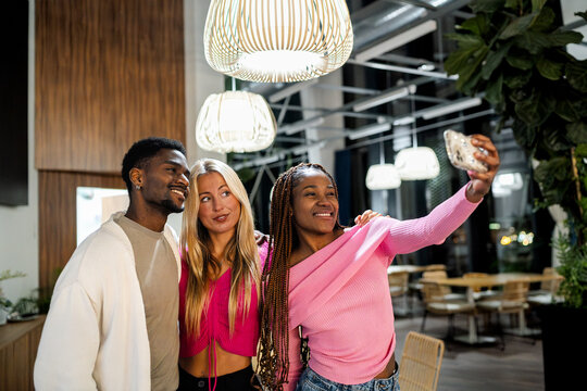 Diverse young adult friends smiling, taking a selfie with a smartphone, and enjoying a night out at a modern restaurant