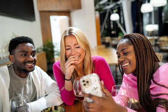 Diverse friends sharing a happy moment at a restaurant, looking at a smartphone and laughing together. Social media connection