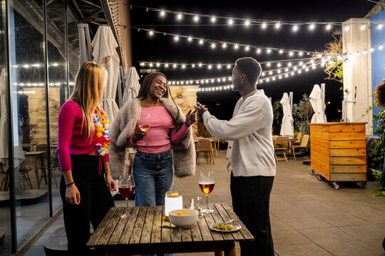 Group of diverse friends socializing and laughing together at an outdoor restaurant or bar, celebrating friendship under string lights