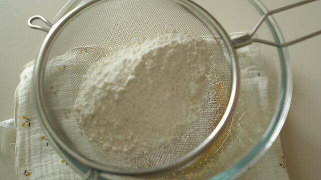 Pouring flour into a sieve over a glass bowl of egg mixture