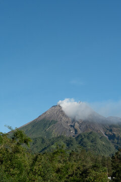 Mount Merapi's broken crater and rocky peak rise above green hills as a faint trail of white smoke escapes from the summit
