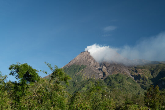 Mount Merapi rises above lush green surroundings with a small white smoke plume emerging from its summit and a weathered crater formed by past eruptions