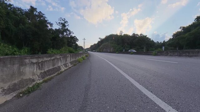 Wide asphalt road curving between forested hills, with concrete roadside barriers, distant traffic and soft golden clouds across a blue evening sky.