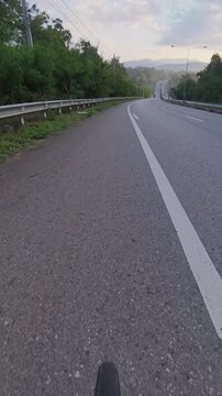 Vertical cycling POV footage of a rolling asphalt road leading toward distant forested mountains, with metal guardrails, white road markings and lush green roadside vegetation under a cloudy sky.