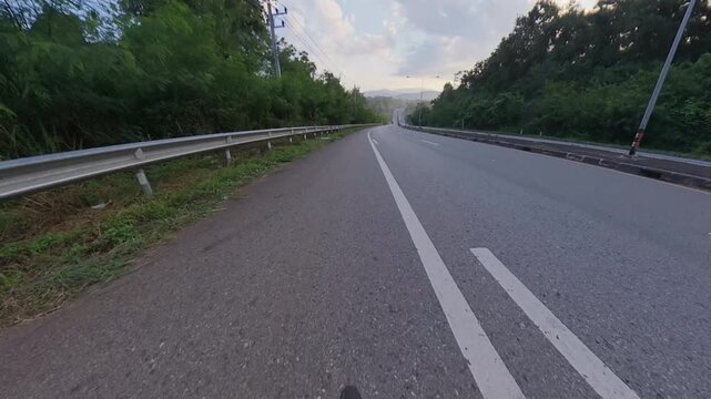Horizontal cycling POV footage descending a wide mountain road toward distant forested hills, with metal guardrails, white lane markings, a steep slope warning sign and lush roadside vegetation.