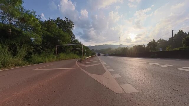 Rural highway leading toward distant mountains at golden hour, with sunlight reflecting on the asphalt, road markings, roadside greenery and passing vehicles beneath a partly cloudy sky.