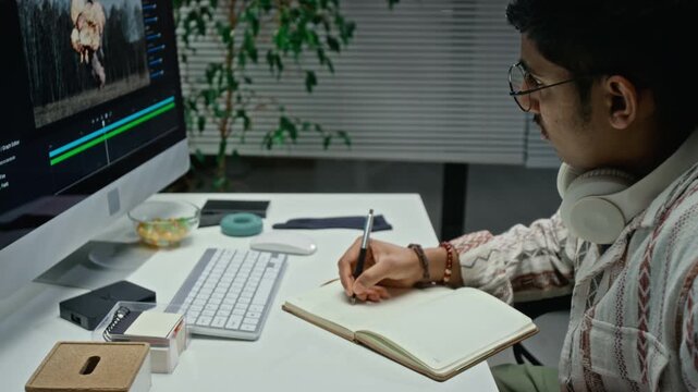 Asian young adult video editor writing notes in notebook next to computer screen during post production process. Footage suitable for multimedia production, digital agency, and creative industry