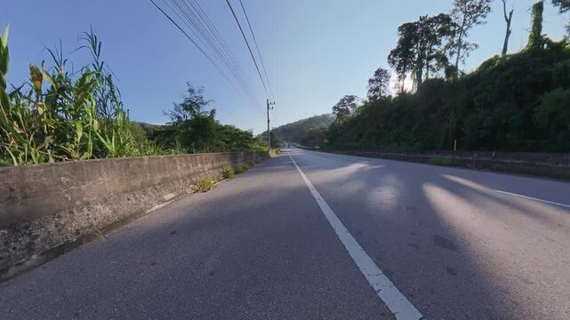 Horizontal cycling POV footage along a quiet rural asphalt road toward green hills, with concrete roadside walls, overhead power lines and low evening sunlight casting long shadows across the pavement