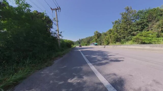 Wide downhill road through forested mountain scenery, with concrete roadside barriers, lush green hills and a clear blue sky on a sunny day.