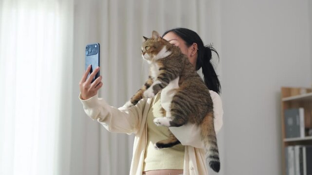 Asian woman taking selfie with pet cat at home, smiling and holding tabby cat in bright living room with smartphone