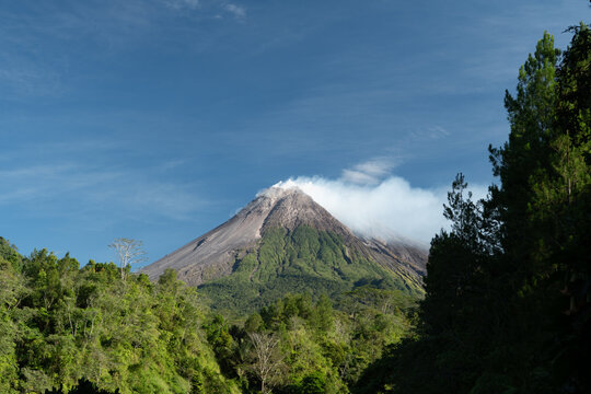 Scenic view of Mount Merapi showing a broken volcanic crater, faint white smoke above the peak and dense green vegetation across the surrounding landscape