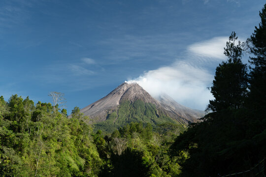 Mount Merapi's rugged volcanic summit releases a faint trail of white smoke above green tropical hills, revealing a crater shaped by historical eruptions
