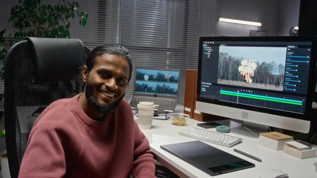 Portrait of Indian young adult visual effects artist editing CGI explosion on computer monitor, turning and smiling. Footage ideal for video production tutorials and tech career advertising
