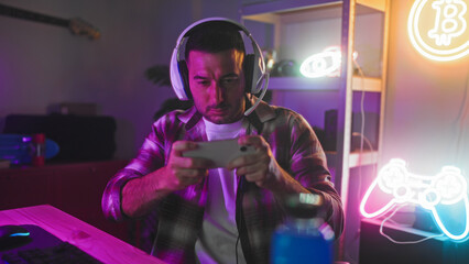 Man holding smartphone with both hands, wearing headset at neon lit streaming studio desk with keyboard, mouse and gamepad sign  concentration challenge. © Krakenimages.com