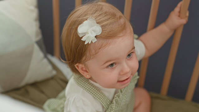 Blonde toddler girl gripping crib rail with hand and smiling face in building nursery; innocent curiosity.