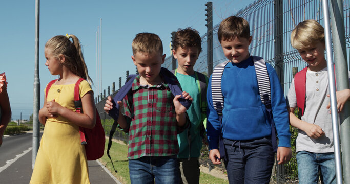 Happy caucasian elementary school friends with schoolbags walking on sunny road to school