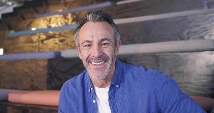 Mature man sitting on tiered bench in lounge, smiling and leaning forward wearing blue shirt