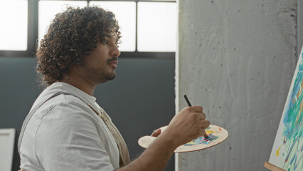 Man painter wearing apron holds palette and brush and paints bold strokes on canvas in studio  focused concentration. © Krakenimages.com