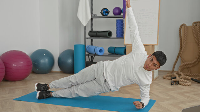 Man doing side plank with raised arm on blue exercise mat in bright fitness studio, wearing white top and gray sweatpants; focus balance training.