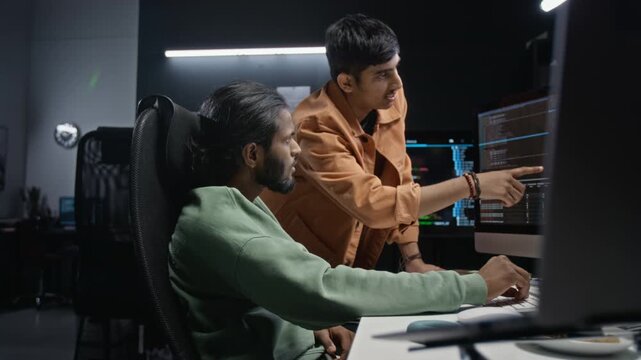Young adult male developers analyzing code on computer monitor in IT office. Ideal for tech startup commercials, programming training, software engineering campaigns