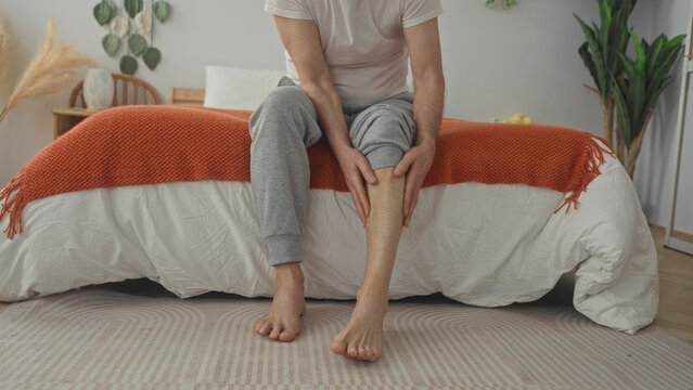 Man middle aged sitting on bed massaging calf for cramp while barefoot and wincing in building; recovery pain.