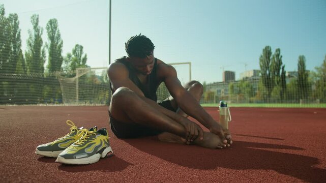 African American adult man male footballer pressing painful toe on urban pitch after workout sportsman callus bone strain injury trauma accident sore foot athlete recovery outdoors medical treatment