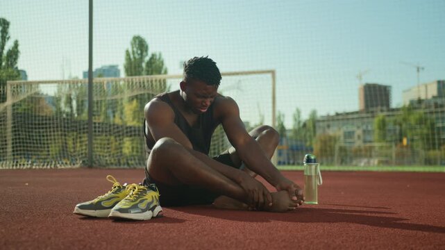 African American man male runner limping after stadium jog rubbing sore calf and blistered foot sports trauma muscle strain painful callus recovery sportsman athlete fitness workout outdoors routine