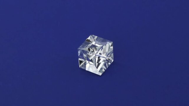 Clear transparent crystal cube rotates continuously on a vibrant dark blue surface, showing intricate geometric facets and light refraction from every single angle