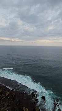 Atmospheric coastal vista of the rugged limestone cliffside, dark ocean waters, and dramatic sky during sunset on the Bukit Peninsula in Bali.