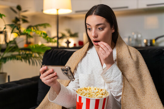 Young woman at night home watches horror on smartphone hugs blanket flinches feels fear so quick breath. Caucasian girl on sofa at evening startles at jump scare tenses grips cup drops popcorn a bit.